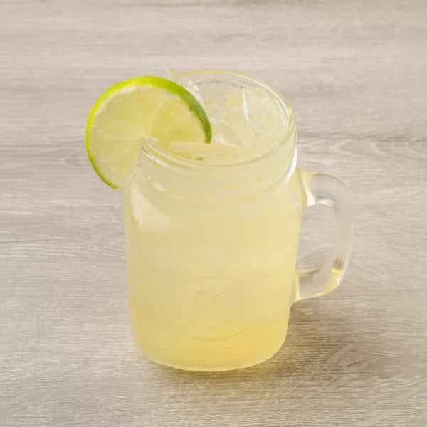 A glass mason jar mug filled with lemonade, ice, and a lime wedge on a wooden table.