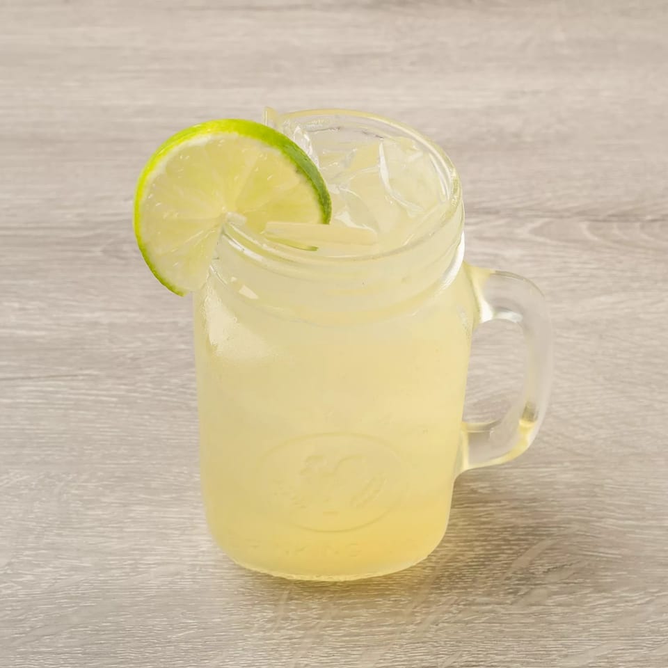 A glass mason jar mug filled with lemonade, ice, and a lime wedge on a wooden table.